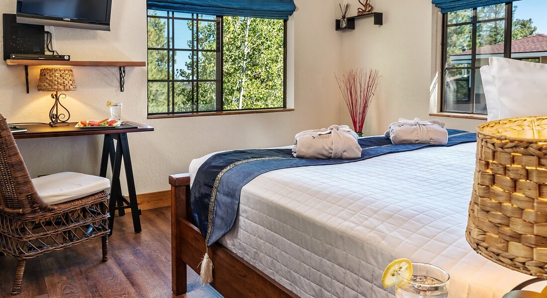 King bed with white comforter and sitting desk with TV on the wall next to large bright windows