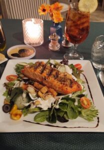 white plate of a salad with dressing and a piece of grilled salmon on a table with ice tea, flowers and a candle