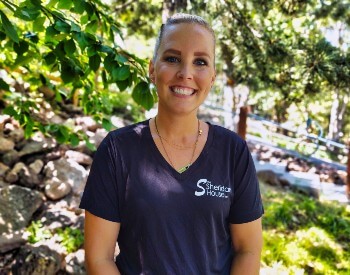 Woman with blond hair pulled back from face, in a black v-neck t-shirt smiling in a wooded area