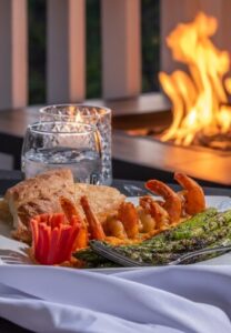 a fire in the background with a table with a water glass and a plate with bread, shrimp and asparagus