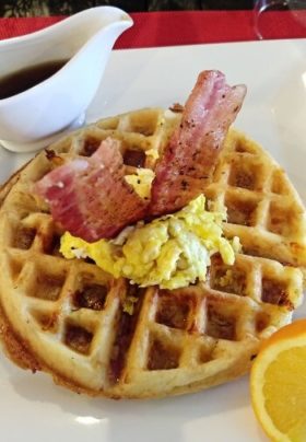 waffle with scrambled eggs and bacon on top with syrup on side