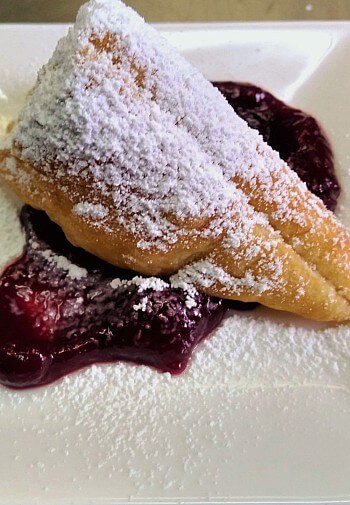 Fried pastry dessert dusted with powdered sugar over a puddle of red sauce on a white plate
