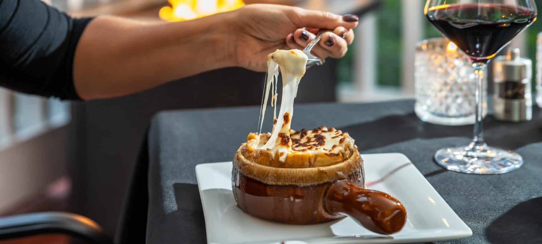 Woman's arm at a table holding a spoon high with melted cheese from a crock of soup