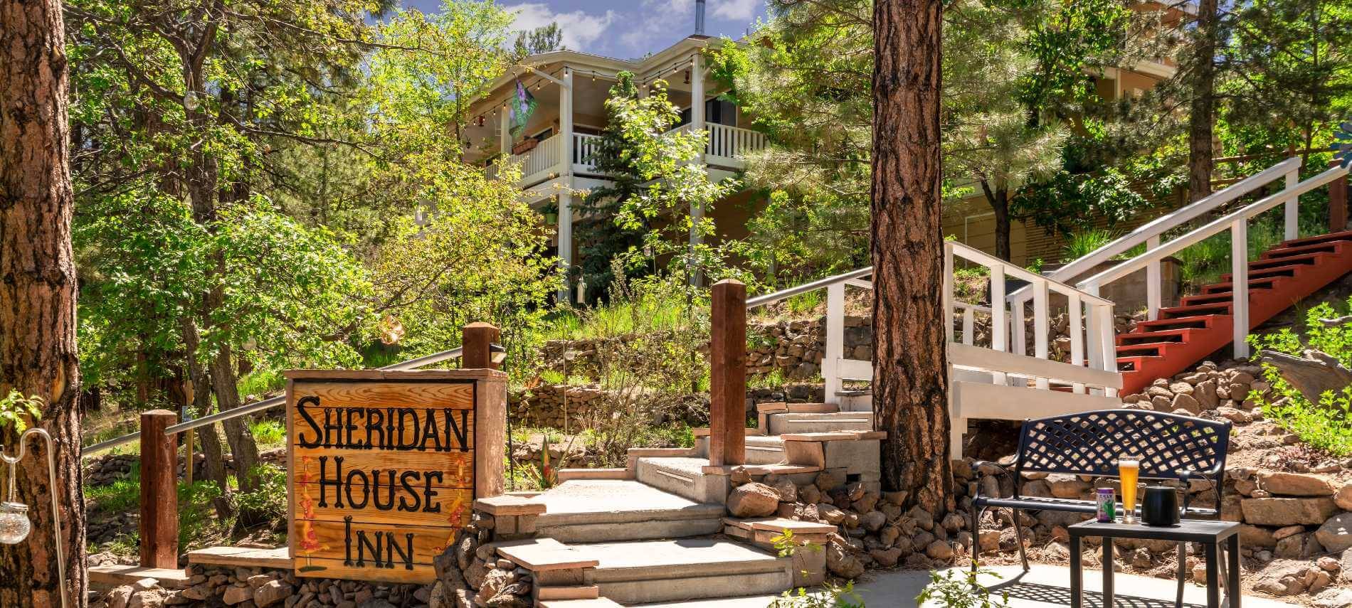 big brown sign with Sheridan House Inn and stairway leading up to a 2 story building with lots of trees and pine tree trunks, with a concrete patio with a bench with table