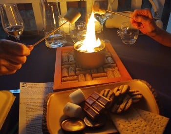 2 people with sticks roasting marshmallows over a small flame with a wood bowl with marshmallows, graham crackers, chocolates and cookies