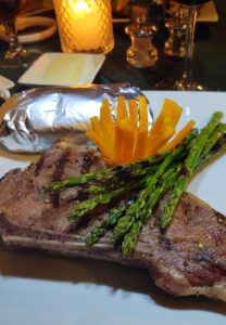 white plate on a table with a candle of a steak, asparagus, potato and a orange bell pepper flower