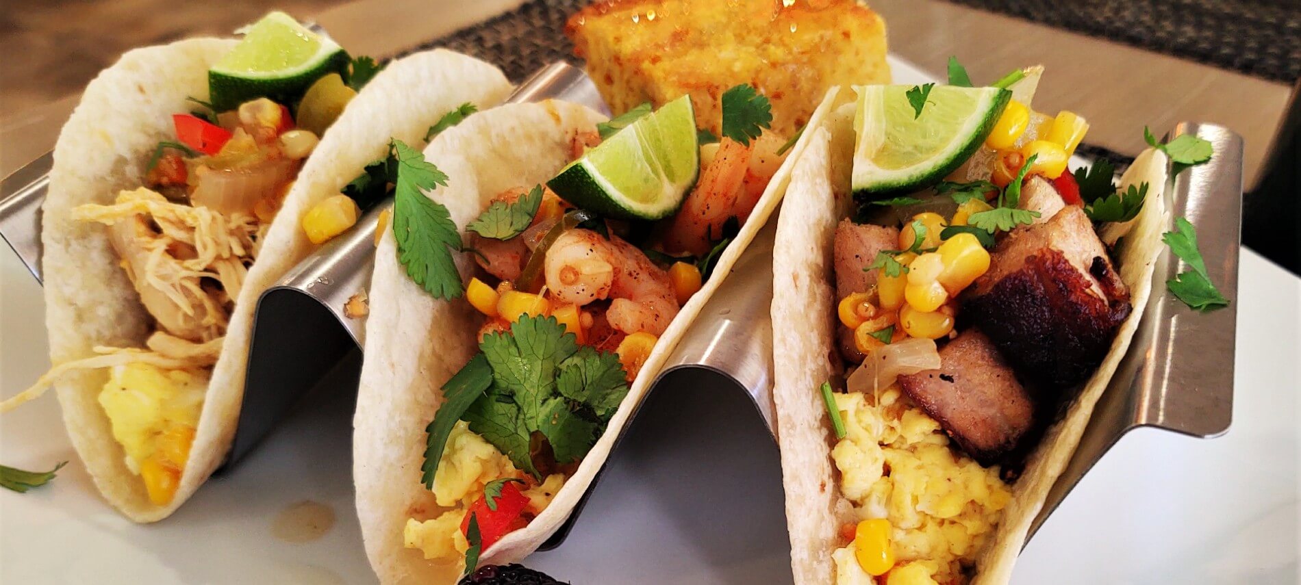 white plate with 3 taco shells villed with a variety of foods, all different, and topped with cilantro and a lime wedge
