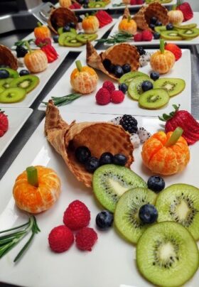 Thanksgiving-themed fruit display on a white porcelain plate featuring a waffle cone shaped like a cornucopia, spilling out blueberries, kiwi, and raspberries, with peeled clementines and celery stems arranged as mini pumpkins, and other fruits scattered decoratively around.
