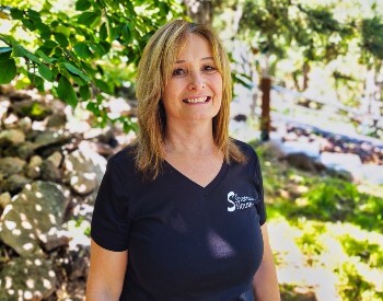 Woman with long light brown hair in a black v-neck t-shirt stands smiling in a wooded area
