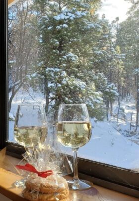 Two glasses of white wine and a wrapped package of Sheridan House Inn’s signature peanut butter welcome cookies on a guestroom windowsill, overlooking snow-covered pine trees in Williams, Arizona.